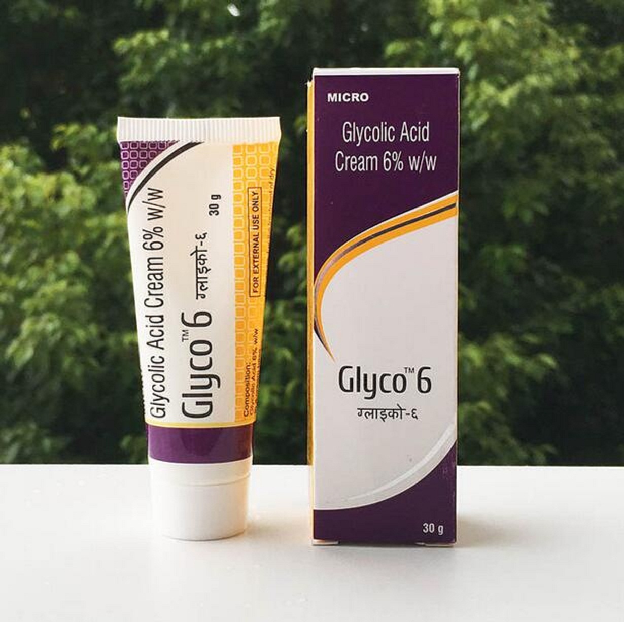Tube and box of Glyco 6 glycolic acid cream displayed on a white surface with greenery behind.