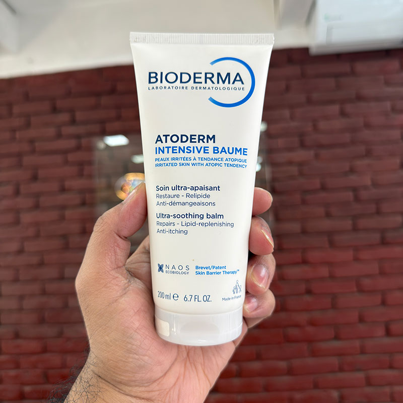 Hand holding a tube of Bioderma Atoderm Intensive Baume in front of a red brick wall.