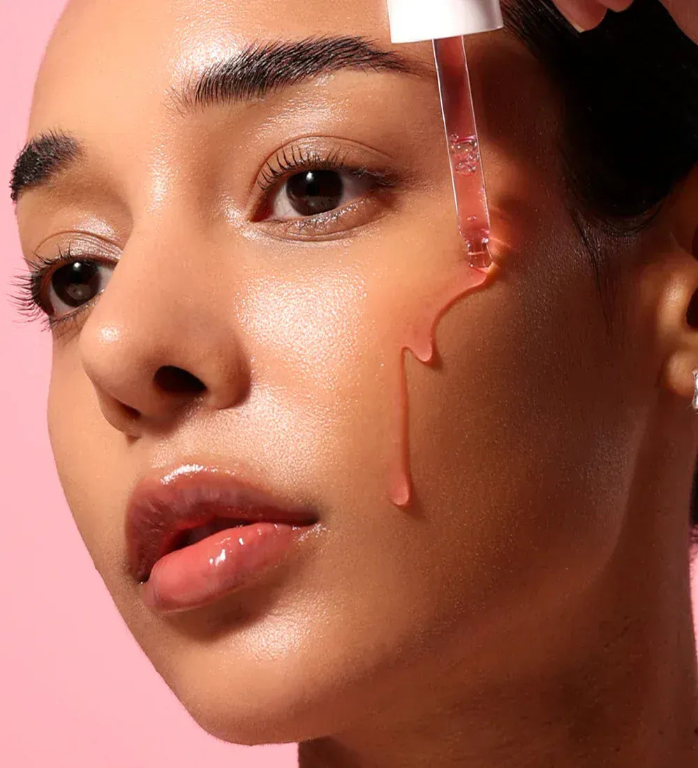 Woman applying serum to her cheek with a dropper against a pink background.