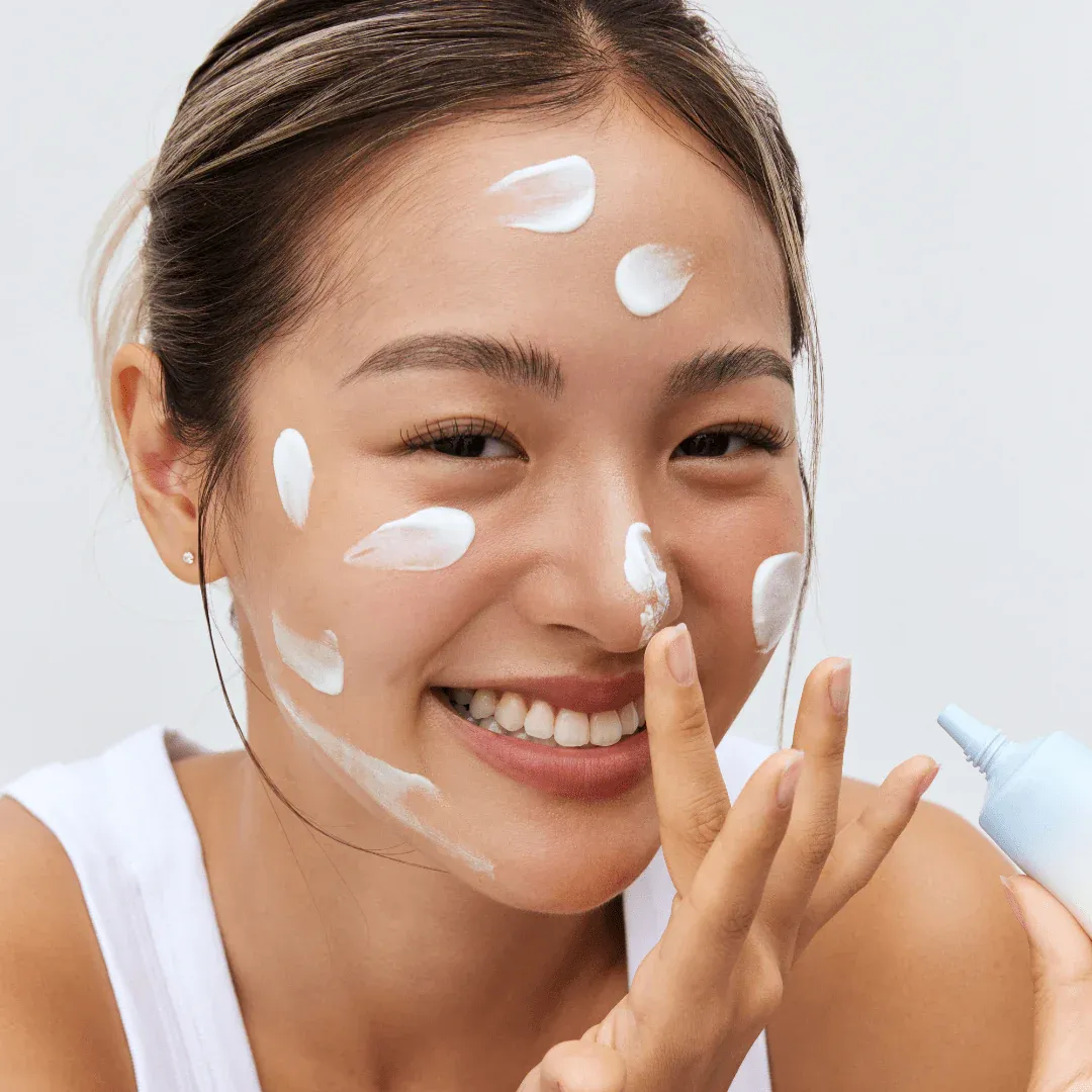 Smiling woman applying white cream to her face, wearing a white top.