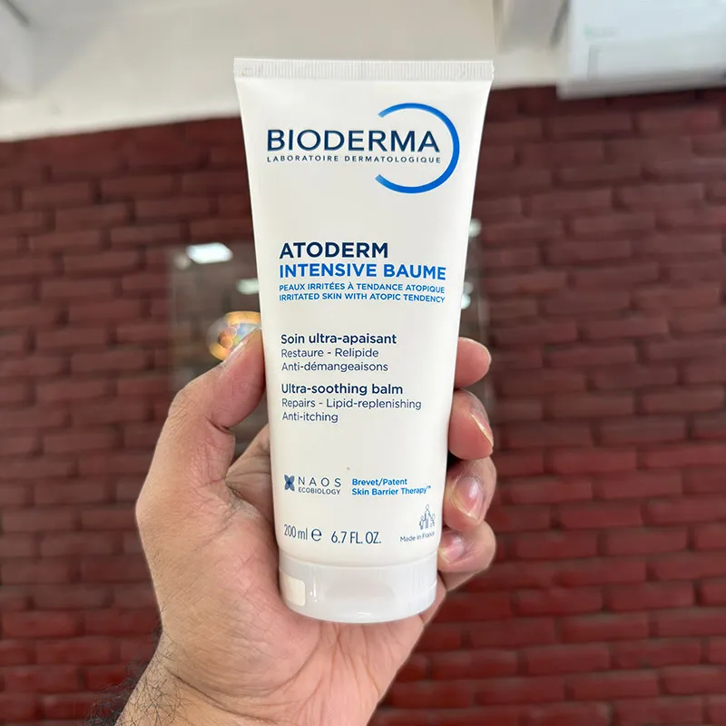 Hand holding a tube of Bioderma Atoderm Intensive Baume in front of a red brick wall.