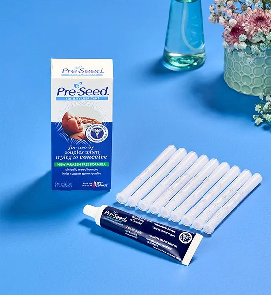 Pre-Seed lubricant box, tube, and applicators on a blue surface with flowers in the background.