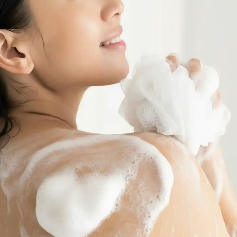 Woman washing shoulder with foamy bath sponge, smiling in a bright setting.